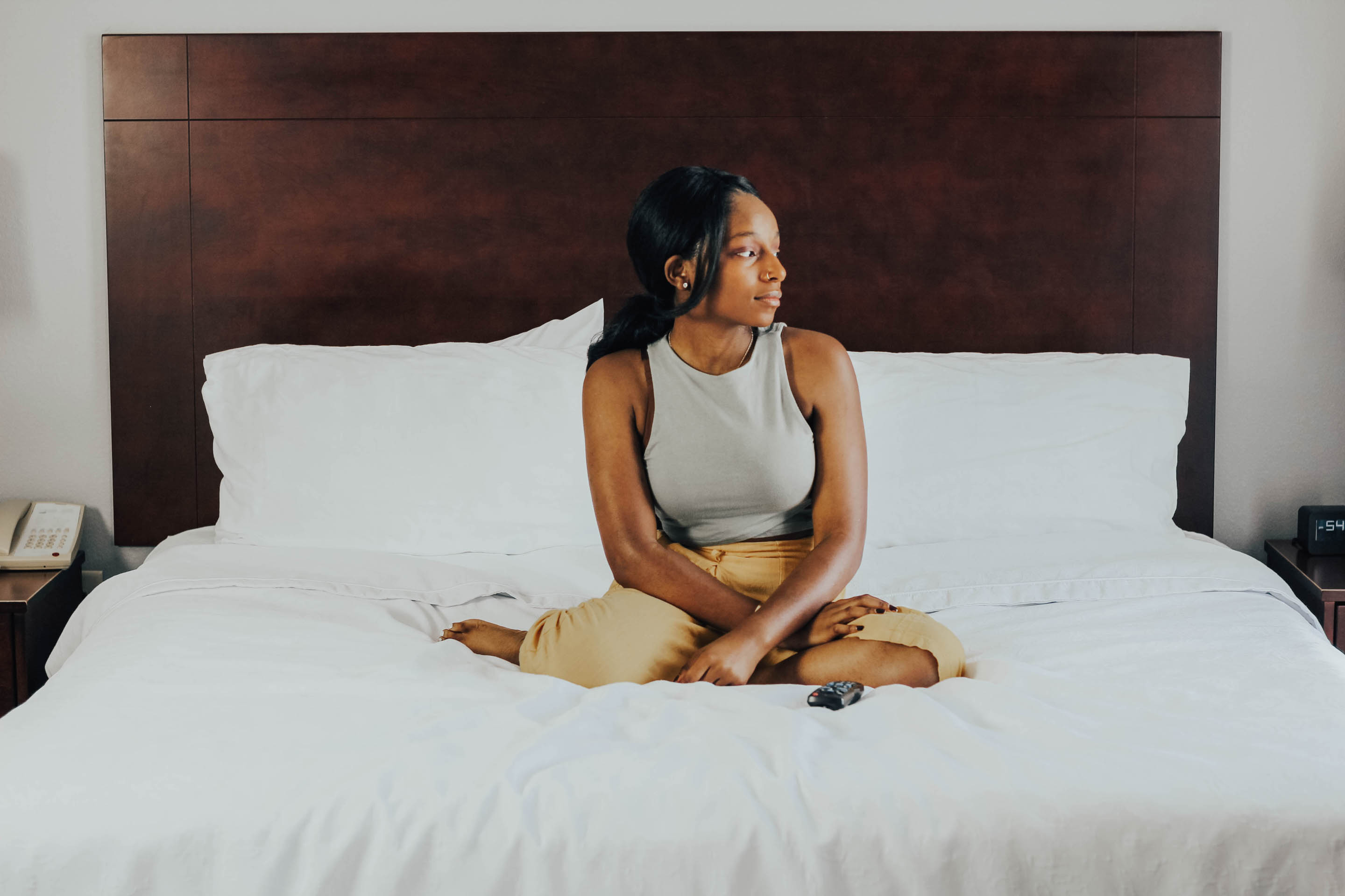 Photo of African American woman dressed in gray tank and tan pants, seated on a kingsized white bed with a dark brown wall-mounted wooden headboard.