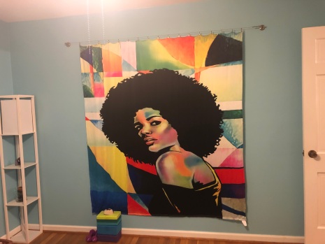 70"x70" fabric tapestry with image of an African American woman wiht a large Afro on a multicolored abstract geometric background. Tapestry is hanging against a blue wall. Two yoga blocks are stacked on the floor.