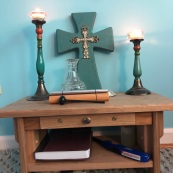 Small wooden seated meditation altar with two lit candles on either end, a teal wooden cross in the center, a small clear glass vase and a chime/mallet. Lower shelf of table has a brown bible and a blue lighter.